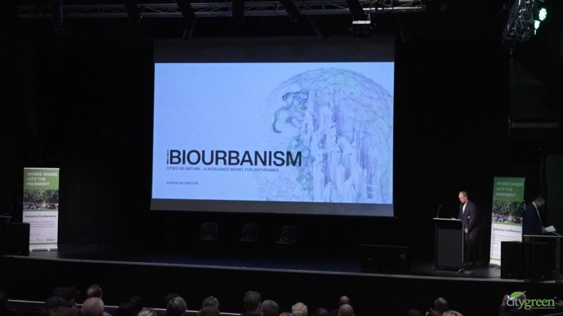 A presentation in a dark auditorium, titled "Biourbanism: Cities as Nature's Resilience Model for Humankind." Author Adrian McGregor stands at a podium in front of the screen with the title. The audience is visible at the bottom of the image, eagerly awaiting insights on biourbanism.